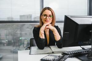 bonito, legal, bonitinho, perfeito mulher sentado às dela escrivaninha em couro cadeira dentro trabalhos estação, vestindo copos, roupa formal, tendo computador portátil e caderno em a mesa foto