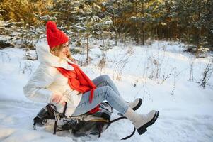 lindo jovem mulher vestindo uma caloroso inverno roupas, sentado em uma trenó e deslizante baixa a colina, tendo Diversão em uma Nevado inverno dia foto