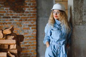 atraente fêmea construção trabalhador dentro capacete. confiante jovem especialista dentro xadrez azul camisa dentro jeans em pé dentro esvaziar sala. interior Projeto e renovação serviço foto