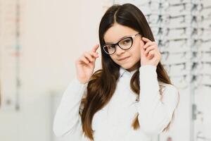 saúde Cuidado, visão e visão conceito - feliz mulher escolhendo óculos às óptica loja. retrato do lindo jovem mulher tentando Novo óculos dentro oculista loja. foto