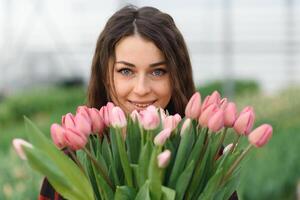 uma retrato do uma fofa jovem mulher com lindo olhos, quem é segurando uma ramalhete do roxa tulipas e representando uma agradável Maquiagem em dela face. foto