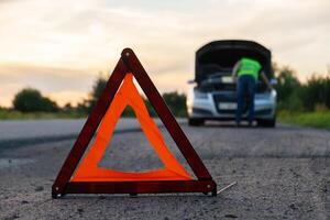 quebrado prata luxo carro emergência acidente. homem motorista instalando vermelho triângulo Pare placa em estrada. esporte automóvel virou em Bloqueios técnico problemas em a estrada. segurança procedimento quando tendo veículo quebrado baixa foto