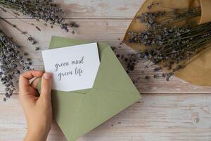 fêmea mãos levando papel cartão Nota com texto verde mente verde vida a partir de envelope. lavanda flor. topo visualizar, plano deitar. conceito do eco sustentabilidade estilo de vida mental espiritual saúde auto Cuidado foto