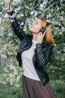 uma ruivo Adolescência menina escuta para música dentro fones de ouvido, configurado através da a telefone. a Tempo do Primavera floração do cereja pomares foto