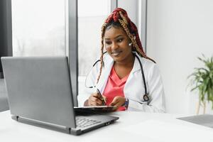 medicamento, conectados serviço e cuidados de saúde conceito - feliz sorridente africano americano fêmea médico ou enfermeira com fone de ouvido e computador portátil tendo conferência ou vídeo ligar às hospital. foto