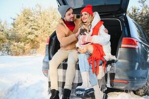 sorridente casal com cachorro sentado dentro aberto suv carro tronco dentro Nevado floresta. desfrutando cada de outros dentro ativo inverno feriados. foto