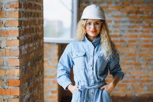 atraente fêmea construção trabalhador dentro capacete. confiante jovem especialista dentro xadrez azul camisa dentro jeans em pé dentro esvaziar sala. interior Projeto e renovação serviço. foto
