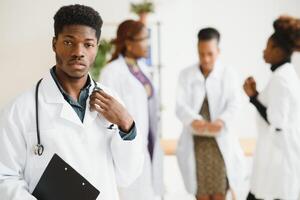 jovem africano masculino médico sorridente enquanto em pé dentro uma hospital corredor com uma diverso grupo do funcionários dentro a fundo. foto