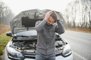 triste motorista segurando dele cabeça tendo motor problema em pé perto quebrado carro em a estrada. carro demolir conceito foto