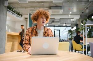 jovem feliz masculino trabalhador autonomo dentro casual roupas sentado dentro cafeteria com computador portátil e usando Móvel telefone. foto