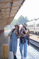 dois jovem ásia amigos meninas com mochilas às estrada de ferro estação esperando para trem, dois lindo mulheres caminhando ao longo plataforma às trem estação foto