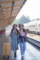 dois jovem ásia amigos meninas com mochilas às estrada de ferro estação esperando para trem, dois lindo mulheres caminhando ao longo plataforma às trem estação foto