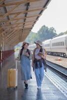 dois jovem ásia amigos meninas com mochilas às estrada de ferro estação esperando para trem, dois lindo mulheres caminhando ao longo plataforma às trem estação foto