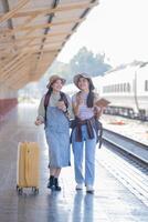 dois jovem ásia amigos meninas com mochilas às estrada de ferro estação esperando para trem, dois lindo mulheres caminhando ao longo plataforma às trem estação foto