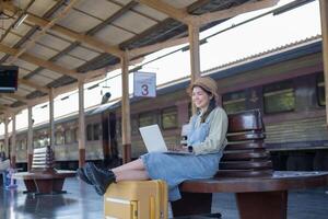 menina usando uma computador portátil enquanto esperando dentro uma trem estação, menina em trem estação com bagagem trabalhando em computador portátil computador, computador portátil dentro usar, senta com uma mala de viagem foto