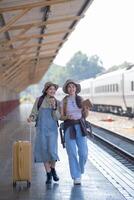 dois jovem ásia amigos meninas com mochilas às estrada de ferro estação esperando para trem, dois lindo mulheres caminhando ao longo plataforma às trem estação foto