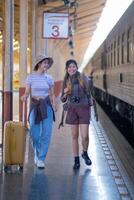 dois jovem ásia amigos meninas com mochilas às estrada de ferro estação esperando para trem, dois lindo mulheres caminhando ao longo plataforma às trem estação foto