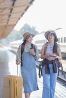 dois jovem ásia amigos meninas com mochilas às estrada de ferro estação esperando para trem, dois lindo mulheres caminhando ao longo plataforma às trem estação foto