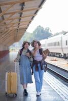 dois jovem ásia amigos meninas com mochilas às estrada de ferro estação esperando para trem, dois lindo mulheres caminhando ao longo plataforma às trem estação foto