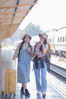 dois jovem ásia amigos meninas com mochilas às estrada de ferro estação esperando para trem, dois lindo mulheres caminhando ao longo plataforma às trem estação foto