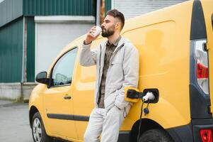 bonito homem bebendo café enquanto cobrando elétrico carro foto