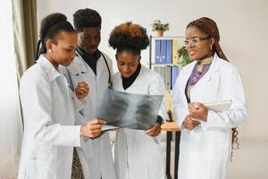 jovem africano masculino médico sorridente enquanto em pé dentro uma hospital corredor com uma diverso grupo do funcionários dentro a fundo. foto