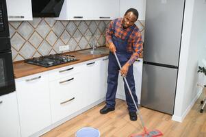 profissional limpador dentro azul uniforme lavando chão e limpando poeira a partir de a mobília dentro a vivo quarto do a apartamento. limpeza serviço conceito foto