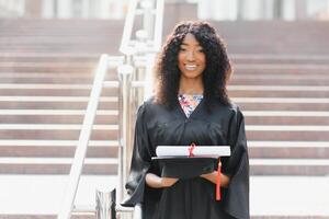 alegre afro americano fêmea graduado em pé dentro frente do universidade construção foto