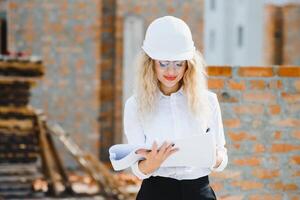 construção engenheiro. menina com construção documentação. uma mulher dentro uma branco Difícil chapéu contra a cobertura do uma prédio. construção do uma Novo casa foto