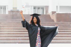 alegre afro americano fêmea graduado em pé dentro frente do universidade construção foto