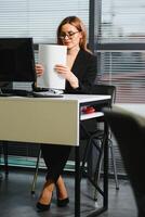 bonito, legal, bonitinho, perfeito mulher sentado às dela escrivaninha em couro cadeira dentro trabalhos estação, vestindo copos, roupa formal, tendo computador portátil e caderno em a mesa foto