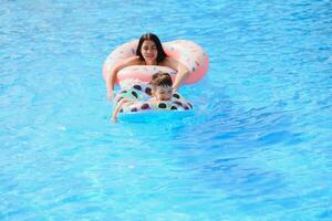 feliz família, jovem ativo mãe e adorável encaracolado pequeno bebê tendo Diversão dentro uma natação piscina, criança Aprendendo para nadar dentro a inflável brinquedo anel, desfrutando verão período de férias às uma tropical recorrer foto