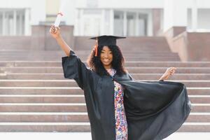 alegre afro americano fêmea graduado em pé dentro frente do universidade construção foto