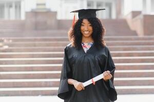 alegre afro americano fêmea graduado em pé dentro frente do universidade construção foto