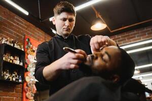 lado Visão do sério homem com à moda moderno corte de cabelo olhando frente dentro barbeiro fazer compras. mão do barbeiro guardando em linha reta navalha e corte na moda listras em cabeça do cliente. foto