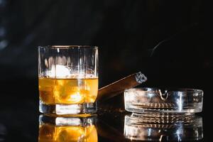 uma vidro do uísque ou bourbon com gelo cubos e uma charuto em uma Preto ardósia com fumaça foto