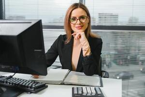 bonito, legal, bonitinho, perfeito mulher sentado às dela escrivaninha em couro cadeira dentro trabalhos estação, vestindo copos, roupa formal, tendo computador portátil e caderno em a mesa foto