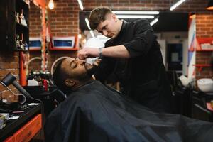 lado Visão do sério homem com à moda moderno corte de cabelo olhando frente dentro barbeiro fazer compras. mão do barbeiro guardando em linha reta navalha e corte na moda listras em cabeça do cliente. foto