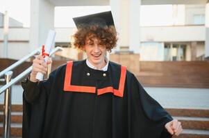 baixo ângulo retrato do feliz triunfante masculino graduado em pé perto universidade segurando acima diploma. a partir de abaixo do jovem bonito homem orgulhoso do acadêmico conquistas a comemorar Faculdade graduação foto