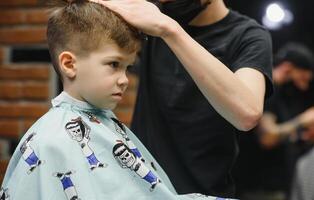 uma pequeno fofa Garoto senta dentro uma cabeleireiro às a estilista, uma aluno é obtendo cabelo cortar dentro uma beleza salão, uma criança às uma barbearia, uma curto masculino corte de cabelo foto