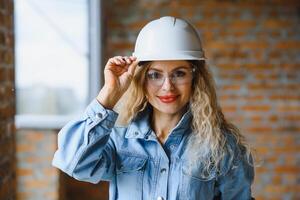 atraente fêmea construção trabalhador dentro capacete. confiante jovem especialista dentro xadrez azul camisa dentro jeans em pé dentro esvaziar sala. interior Projeto e renovação serviço. foto