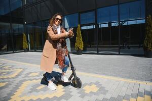 jovem lindo mulher dentro uma Jaqueta sorrisos e passeios a elétrico lambreta para trabalhos ao longo escritório edifícios foto