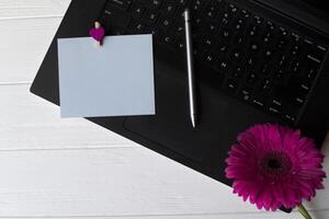 Preto computador portátil teclado, azul esvaziar papel e roxa gerbera em uma branco de madeira trabalhos escrivaninha. foto