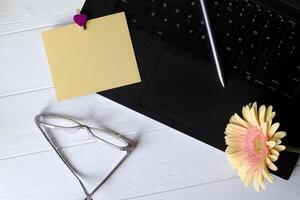 Preto computador portátil teclado, amarelo esvaziar papel, óculos e bege gerbera em uma branco de madeira trabalhos escrivaninha. fêmea ambiente de trabalho. foto
