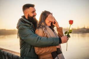 feliz homem dando vermelho rosa para dele mulher enquanto elas apreciar gastos Tempo juntos em uma pôr do sol. foto