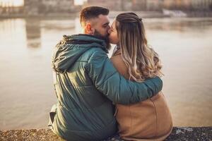 feliz casal se beijando e abraçando enquanto apreciar sentado de a rio. foto