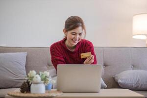 conectados compras. mulher comprando computador portátil usando crédito cartão sentado em sofá às casa foto