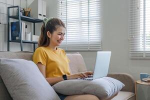 lindo ásia mulher vestindo casual roupas em a sofá usando uma computador portátil computador, divertido com social meios de comunicação, relaxante, sorridente e rindo. foto