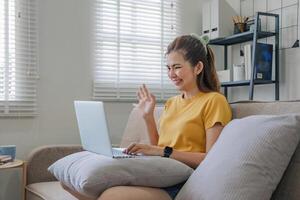 lindo ásia mulher vestindo casual roupas em a sofá usando uma computador portátil computador, divertido com social meios de comunicação, relaxante, sorridente e rindo. foto