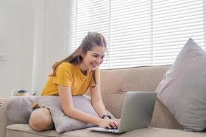 lindo ásia mulher vestindo casual roupas em a sofá usando uma computador portátil computador, divertido com social meios de comunicação, relaxante, sorridente e rindo. foto
